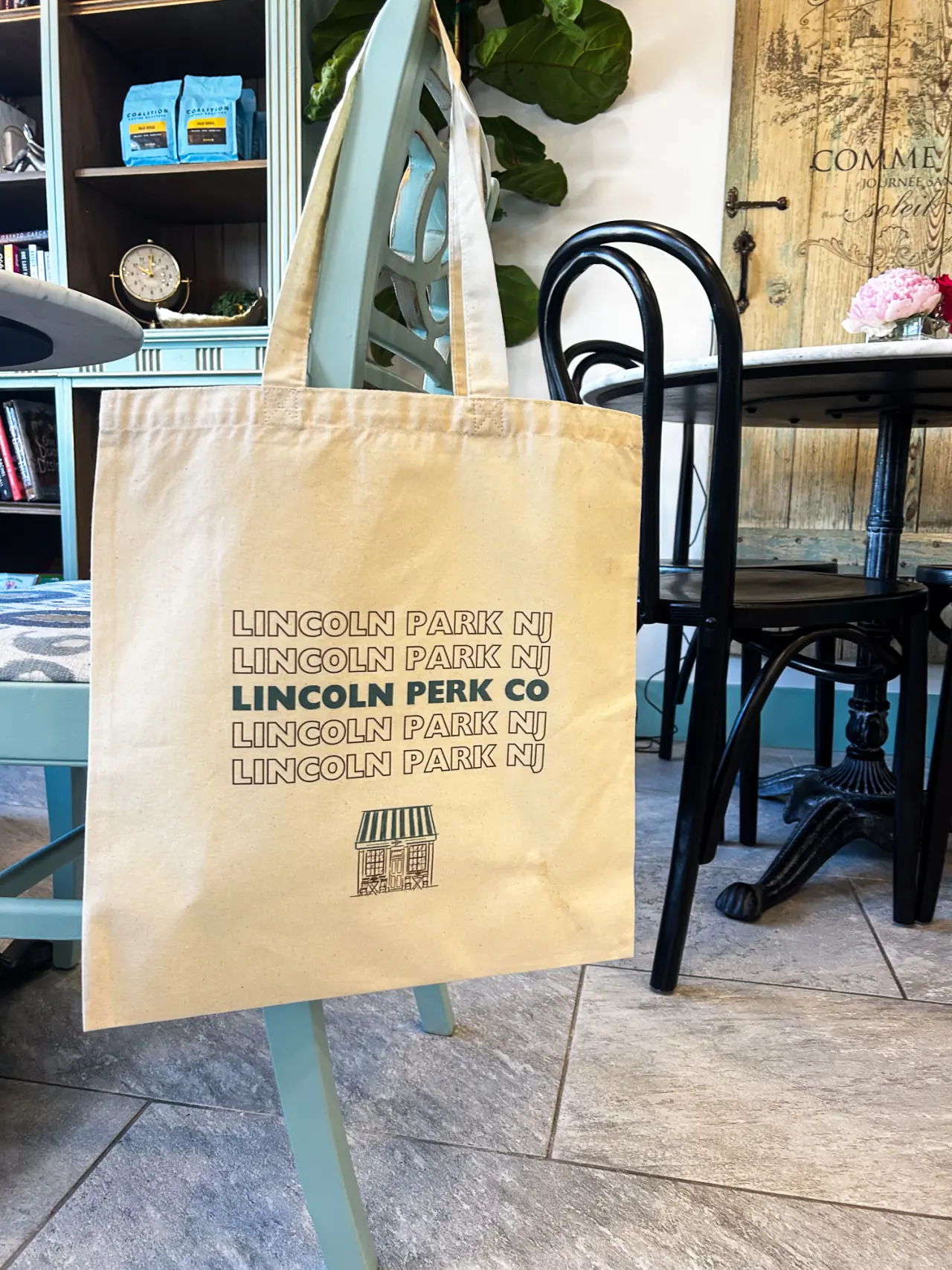 Canvas Lincoln Perk Co tote bag hanging on a chair inside the caf&eacute;, with marble tables, bentwood chairs and shelves of coffee bags behind it