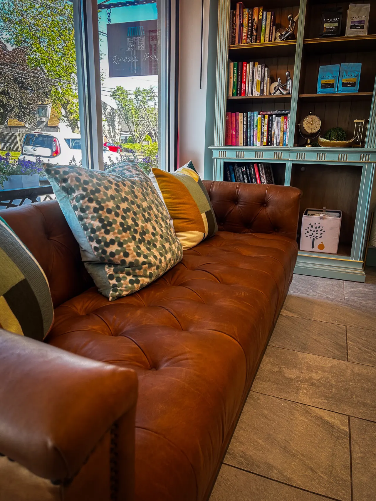 The brown leather chesterfield sofa with patterned pillows, next to the teal bookcase and storefront window